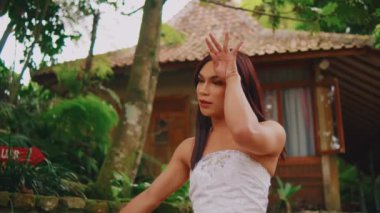 Geleneksel beyaz kembenli güzel bir Asyalı kadın ve batik saçlarını düzeltiyor ve yemyeşil bir bahçe arka planında sakin bir ifadeyle etrafına bakıyor..