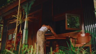 Kırsal bir evin ahşap korkuluklarına yaslanan kişi gün ışığında yemyeşil ile çevrili.