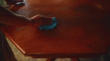 Koyu renk bir ahşap masayı mavi bir mikrofiber bez kullanarak temizleyen bir elin detaylı yakın plan görüntüsü, kırsal bir ortamda iç bakım ve hijyen üzerine odaklanıyor..