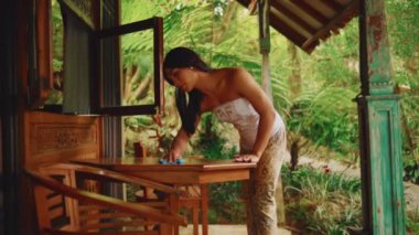 Geleneksel Endonezya elbisesi içindeki güzel bir kadın dikkatle bir orman bahçesinin doğal güzelliğiyle çevrili ahşap bir masayı terasa siler..