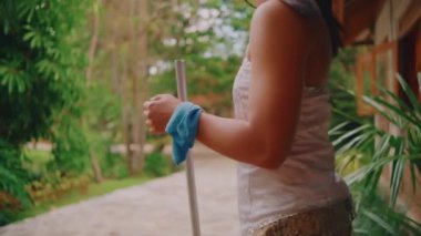 Üzerinde mavi bir bez bulunan süpürge sapını kavrayan bir kadının, bulanık, yemyeşil bir bahçeye ve geleneksel bir eve karşı çekilmiş detaylı bir fotoğrafı..