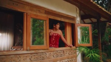 Süslü ahşap bir pencereden sarkan, elinde bir bardak suyla konuşan ve el kol hareketi yapan geleneksel kırmızı elbiseli bir kadının orta boy fotoğrafı..