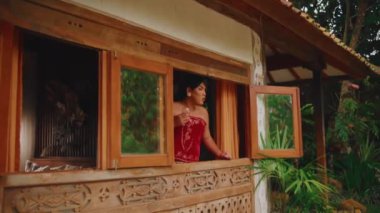 Kırmızı askısız bluz ve batik sarong giyen bir kadın elinde bir bardak suyla, kırsal bir kulübenin ahşap penceresinden etrafına tuhaf bir şekilde bakıyor..