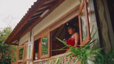 Kırmızı etnik kıyafetli bir kadın, ahşap pencere çerçevesine yaslanarak geleneksel bir köy evinin sakin atmosferinin tadını çıkarırken bir yudum su alır..