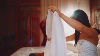 İki kadın bir odada, biri beyaz elbiseyi ayarlıyor, gece boyunca özel bir etkinliğe hazırlanıyor.