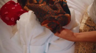 Beyaz çarşaf üzerinde geleneksel bir Java Batik kumaşı düzenleme ve katlama, aile içi bakım ve miras tekstilini temsil eden ellerin yakın plan görüntüsü.
