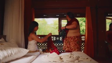 Öfkeli yaşlı bir kadının, geleneksel ahşap bir odada giysilerini toplayıp katlayan genç bir kadını azarladığı gergin bir aile sahnesi..