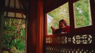 Süslü, ahşap bir pencere çerçevesinin içinden sakin bir manzara, yemyeşil bir bahçeye bakıyor, sabah boyunca huzur ve doğal güzelliği aktarıyor.