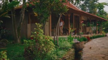 Geleneksel giyinmiş üzgün bir kadının, yemyeşil bir bahçede, taştan bir patikada yavaşça ve yalın ayak yürüdüğü geniş bir kare..