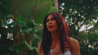 Düşünceli, kızıl saçlı bir kadının portresi. Tropik bir bahçede duruyor. Arka planda büyük yeşil yapraklar var..