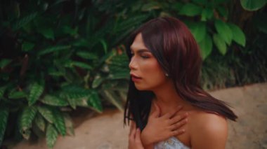 Düşünceli, kızıl saçlı bir kadının portresi. Tropik bir bahçede duruyor. Arka planda büyük yeşil yapraklar var..