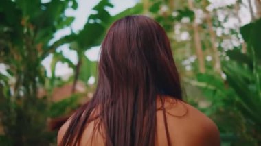 Omuzlarının üstünden, yemyeşil ve ahşap bir çardağı olan tropik bir bahçeye bakan bir kadının manzarası, bir huzur ve doğa ile bağlantı hissi veriyor..