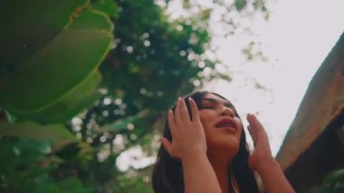 Sıkıntılı genç bir kadın, elleri kulaklarıyla bir bahçede duruyor. Panik, gürültü ya da travmatik anılardan bunalmış bir şekilde, umutsuzluk içinde yukarı bakıyor..