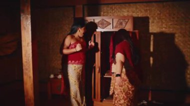 Geleneksel Güney Asya kıyafetli iki kadın kapalı alanda kültürel bir törenle meşgul olurken, bir kadın diğer kıyafetlerini ahşap bir dolabın yanında düzeltiyor..