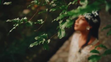 Yeşil yapraklara bakarak sanatsal tepeden tırnağa çekim. Odak, yapraklardan, taş bir yolda duran beyaz elbiseli bir gelinin bulanık figürüne kayıyor..