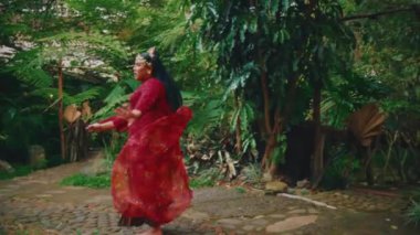 Bahçede dönen, şeffaf kırmızı şalının havada süzülmesine izin veren, geleneksel kıyafetlerini sergileyen bir gelinin büyüleyici yavaş çekim görüntüsü..