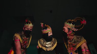 Geleneksel Cava dans kostümleri ve maskeleriyle üç sanatçı, karmaşık başlıklarıyla, loş ışıklandırılmış bir ortamda, sergilenen kültürel sanatlarla yakalandılar..