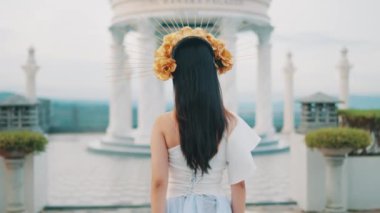 Uzun siyah saçlı, beyaz elbiseli ve altın çiçekli başlıklı bir kadın sırtı kameraya dönük, sütunlu beyaz bir çardağın karşısında duruyor..