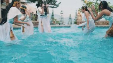 Beyaz elbiseli bir grup kadın ağaçlarla çevrili bir havuzda eğleniyor ve su sıçratıyorlar..