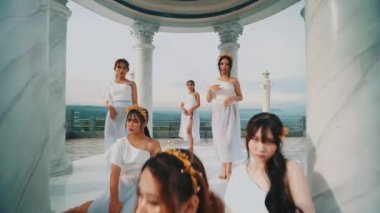 Beyaz giyinmiş bir grup kadın, manzaralı mermer bir balkonun merdivenlerinde zarif bir şekilde poz veriyorlar..