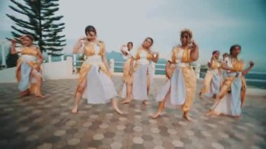 Geleneksel altın ve beyaz kostümlü bir grup kadın, ağaçların ve okyanusun manzaralı arka planına sahip açık bir platformda dans ediyorlar..