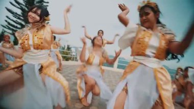 Beyaz ve altın kostümlü bir grup kadın arka planda bulutlu bir gökyüzü ve ağaçlarla döşeli bir zeminde senkronize dans gösterisi yapıyorlar..