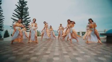 Koordineli altın ve beyaz kostümlü bir grup kadın ağaçlarla kaplı bir yüzey ve arka planda bulutlu bir gökyüzü üzerinde dans gösterisi yapıyorlar..