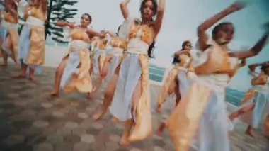 Altın ve beyaz geleneksel giysiler giymiş bir grup kadın açık havada dans ediyorlar. Dağların ve ağaçların manzaralı arka planına sahip fayanslı bir platformdalar.