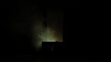 Karanlık, karamsar bir sahne, belli belirsiz bir ışık, bir masa ve bir fon aydınlatıyor. Atmosfer gizemli ve ürkütücü, gölgeler ve duman bir şüphe hissi yaratıyor..