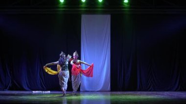 Geleneksel giyinmiş iki dansçı dramatik ışıklandırma ve karanlık bir arka planla sahnede gösteri yapıyorlar..
