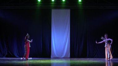 Renkli kostümler içinde iki geleneksel dansçı, loş bir sahnede, beyaz bir zemin ve yukarıda yeşil ışıklar eşliğinde şarkı söylüyorlar..