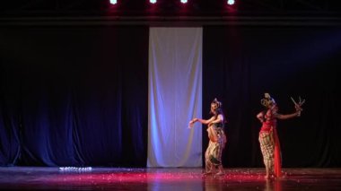 İki dansçı, loş bir sahnede, beyaz bir arka planda geleneksel bir dans gösterisi yapıyorlar. Geleneksel kostümler ve başlıklar giyiyorlar ve sahneyi aydınlatan kırmızı ışıklar var..