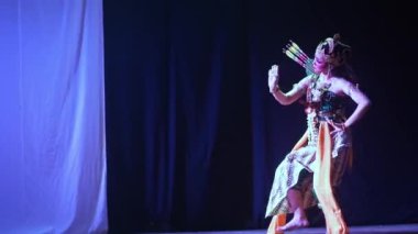 Geleneksel giyinmiş iki dansçı sahnede mavi ve siyah bir arkaplanla gösteri yapıyorlar. Renkli kostümler ve başlıklar giyiyorlar ve bir dansçının sırtında ok titremesi var..