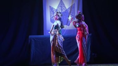 İki geleneksel dansçı sahne arkasında gölge kuklayla gösteri yapıyor. Renkli geleneksel kostümler giyiyorlar ve hareketin ortasındalar..