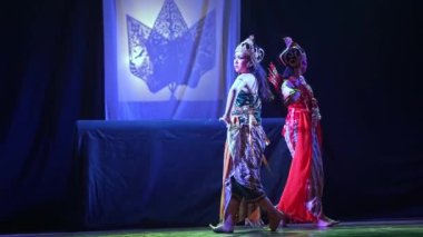 Özenle hazırlanmış kostümlü geleneksel bir dansçı arka planda gölge bir kukla perdesiyle loş bir sahnede performans sergiliyor..
