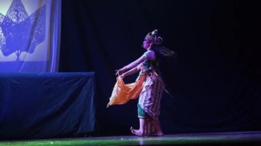 Özenle hazırlanmış bir kostüm giymiş geleneksel bir dansçı sahne arkasında koyu bir fon ve arka planda yansıtılan bir görüntüyle sahnede performans sergiliyor..