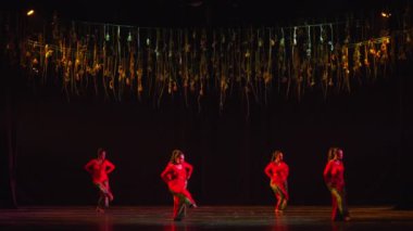 Kırmızı elbiseli dansçılarla modern dans performansı dramatik ışıklandırma altında karanlık bir sahnede asılı dekorasyonlarla.