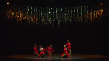 Kırmızı kostümlü oyuncular, karanlık bir sahnede asılı duran, dramatik bir dans ya da tiyatro gösterisi sergileyen.