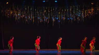 Sahnedeki dansçılar, arka planda dramatik ışıklandırma ve asılı aksesuarlarla çağdaş bir gösteri sergiliyorlar..