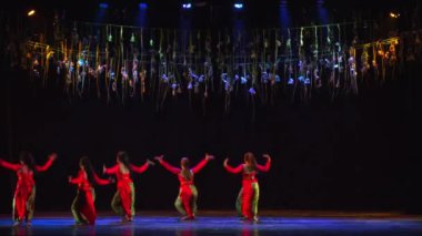 Sahnedeki dansçılar altın flamalar altında gün ışığında dramatik ışıklandırmalarla performans sergiliyorlar.