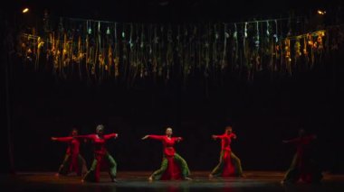 Sahnede dramatik ışıklandırma ve karanlık bir arka planla dans eden dansçılar, gün ışığında asılı duran altın aksesuarlar altında gösteri yapıyorlar.