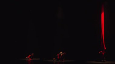 Sahnede kırmızı ışıklandırmalı siluetli dansçılar karanlık bir tiyatro ortamında çağdaş bir dans gösterisi sergiliyorlar..