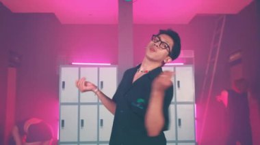 Gözlüklü ve beyaz pantolonlu biri dolapların önünde iki kişiyle birlikte pembe ışığın altında dans ediyor..