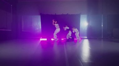 Beyaz elbiseli bir kadın, mor ışıklı bir stüdyoda enerjik bir şekilde dans ediyor..