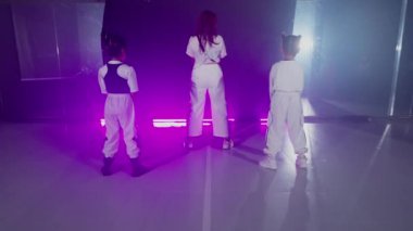 Üç kişi, bir yetişkin ve iki çocuk, beyaz kıyafetler içinde, sırtı kameraya dönük bir şekilde mor ışıklı bir odada duruyorlar..