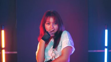 Stüdyoda renkli ışıklarla dans eden kızıl saçlı bir kadın. Beyaz bir elbise ve siyah bileklik takıyor. Arka plan iki dikey ışık, biri turuncu ve biri mavi..