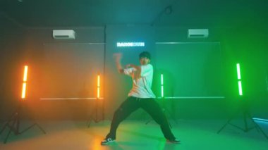 Renkli ışıklandırmayla stüdyoda dans eden biri. Stüdyoda turuncu ve yeşil renkli neon ışıklar var ve kişi dinamik bir dans hareketi yapıyor..