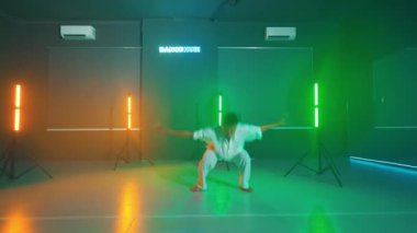 Renkli LED ışıkları olan bir stüdyoda dövüş sanatları pratiği yapan biri. Beyaz dövüş sanatları üniforması giyiyor ve kameradan uzak duruyor. Stüdyo, neon ışıkları ve aynaları olan modern bir tasarıma sahip..