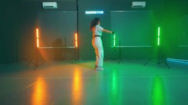 Beyaz elbiseli bir kişi renkli ışıklarla bir stüdyoda dans ediyor. Stüdyonun, turuncu ve yeşil renkler saçan, neon ışıklı, koyu bir arka planı vardır..