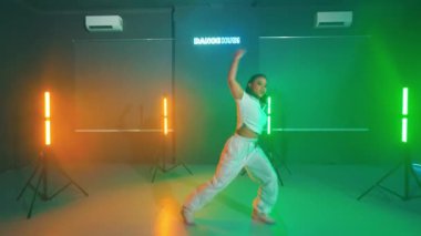 Stüdyoda renkli ışıklarla dans eden bir kadın. Beyaz bir bluz ve beyaz pantolon giyiyor. Stüdyoda neon ışıklar ve duvarda bir tabela var..